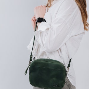 May include: A dark green leather crossbody bag with a silver zipper and a small logo on the front. The bag is worn by a person wearing a white shirt and gray jeans.