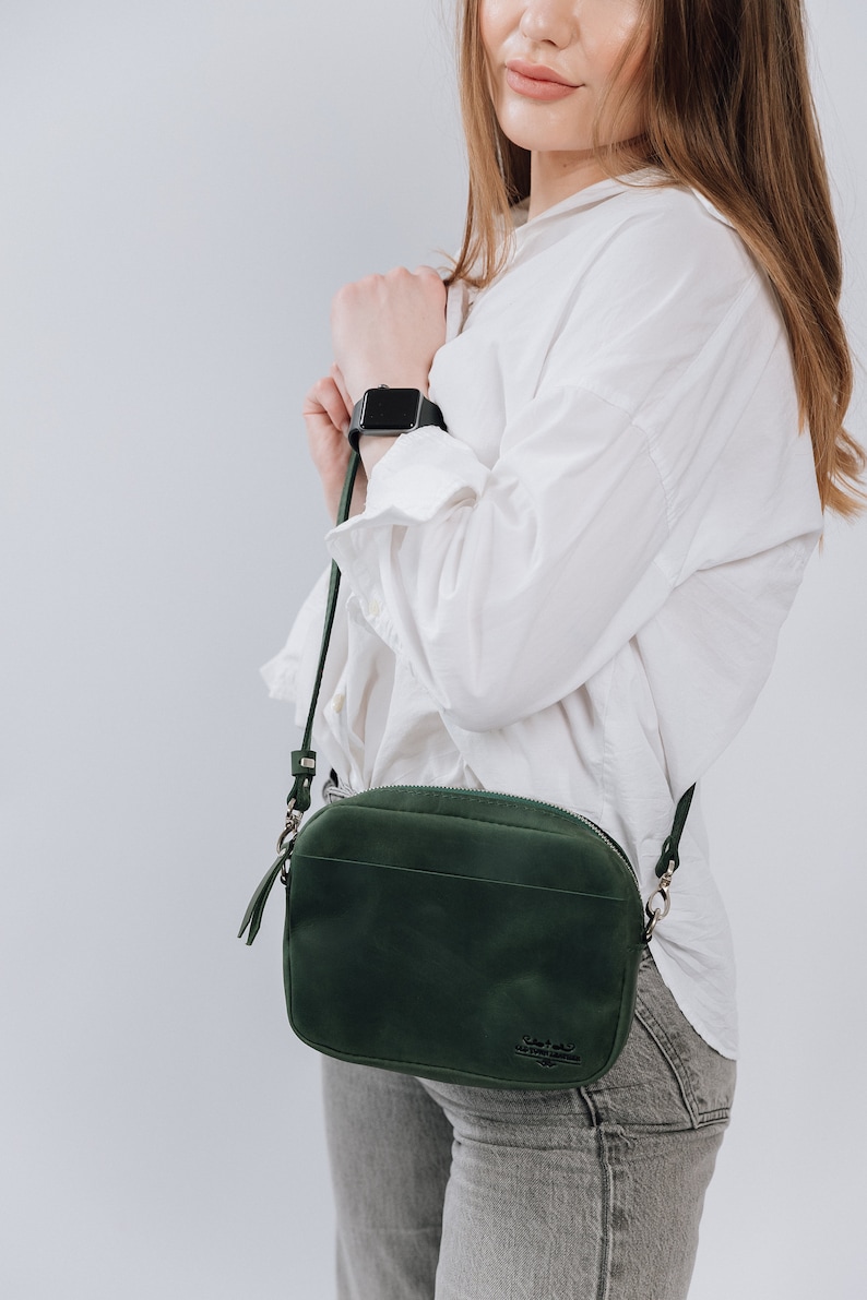 May include: A dark green leather crossbody bag with a silver zipper and a small logo on the front. The bag is worn by a person wearing a white shirt and gray jeans.