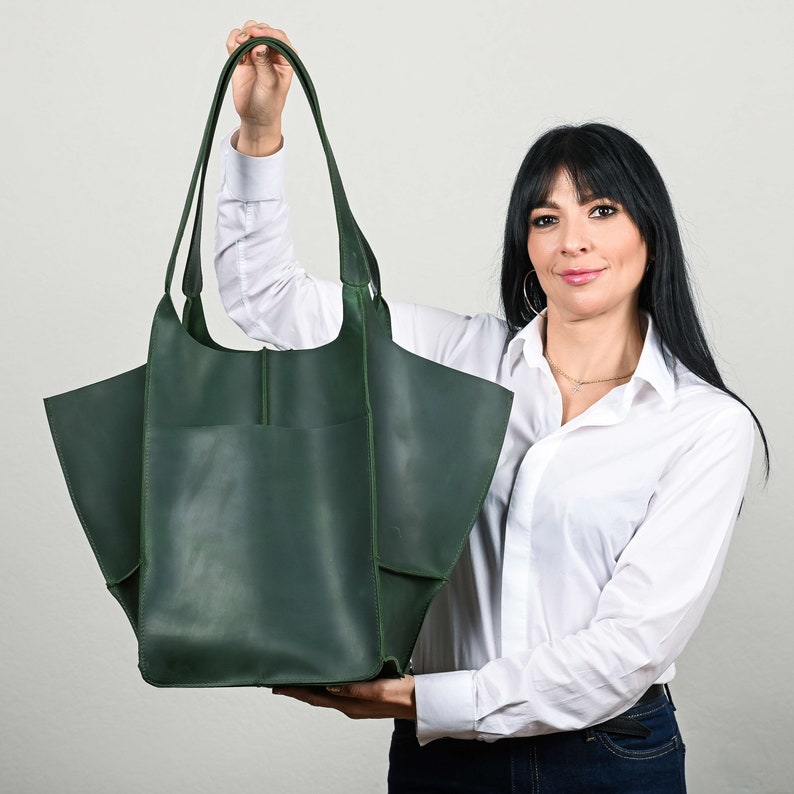 Puede incluir: Un bolso tote de cuero verde oscuro con asas largas y parte superior abierta. El bolso tiene un bolsillo frontal grande y un dise&ntilde;o angular &uacute;nico. El bolso es sostenido por una persona. El bolso mide aproximadamente 38 cm de ancho y 36 cm de alto.