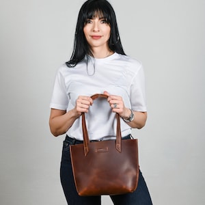 May include: A brown leather tote bag with a top handle. The bag is being held by a person wearing a white t-shirt and blue jeans.