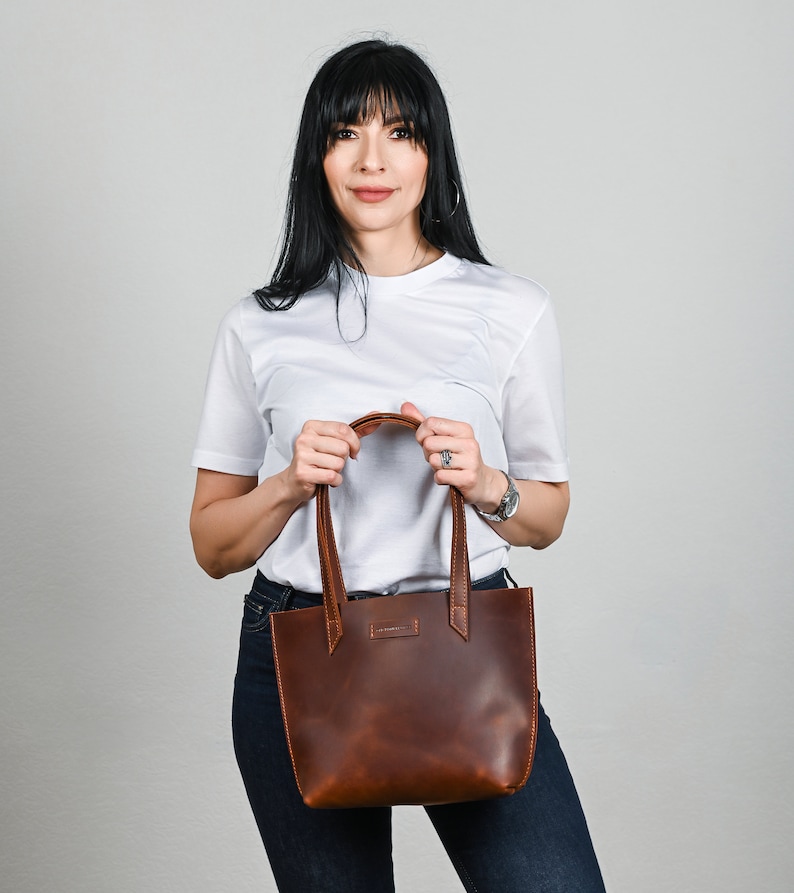 May include: A brown leather tote bag with a top handle. The bag is being held by a person wearing a white t-shirt and blue jeans.