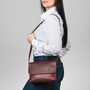 May include: A deep burgundy leather crossbody bag with a rectangular flap closure and a thin, adjustable shoulder strap. The bag features a smooth, matte finish and is worn at the hip, showcasing a clean, structured design with minimal hardware.