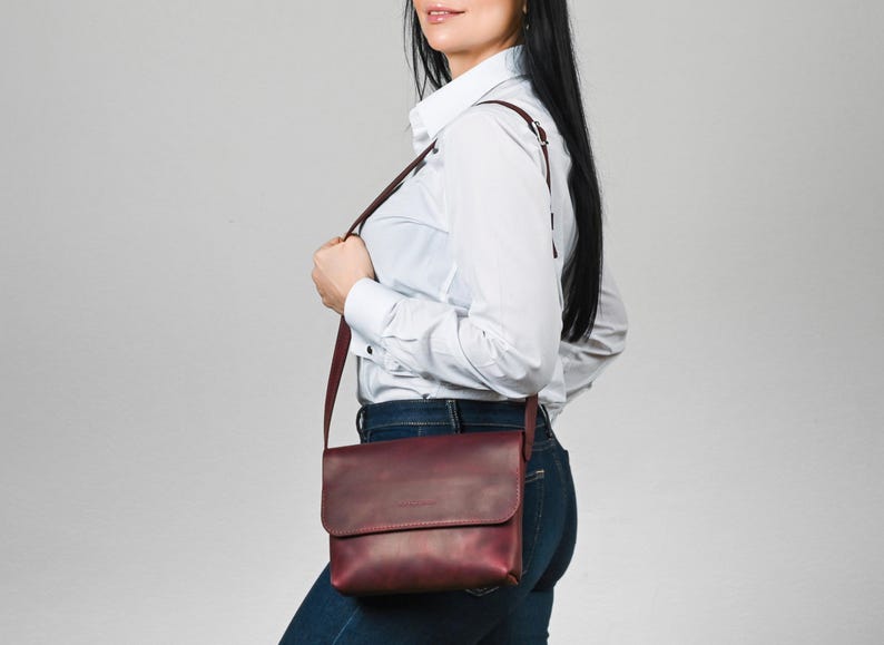 May include: A deep burgundy leather crossbody bag with a rectangular flap closure and a thin, adjustable shoulder strap. The bag features a smooth, matte finish and is worn at the hip, showcasing a clean, structured design with minimal hardware.