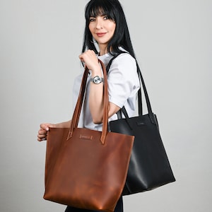 May include: A woman holds two leather tote bags. One is a rich brown, the other is black. Both bags have long handles and a rectangular shape. The brown bag has visible stitching and a small label near the top.