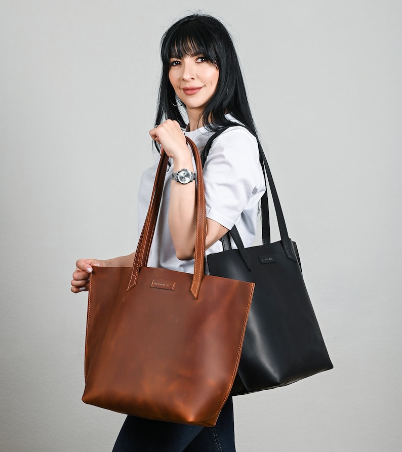 May include: A woman holds two leather tote bags. One is a rich brown, the other is black. Both bags have long handles and a rectangular shape. The brown bag has visible stitching and a small label near the top.