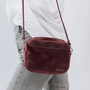 May include: A burgundy leather crossbody bag with a zipper closure and a gold metal logo that reads "Old Town Leather".