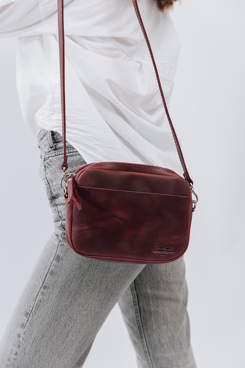 May include: A burgundy leather crossbody bag with a zipper closure and a gold metal logo that reads "Old Town Leather".