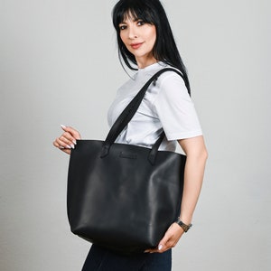 May include: A black leather tote bag with two shoulder straps. The bag is rectangular with a flat bottom and a minimalist design. The bag is held by a person wearing a white t-shirt and blue jeans.