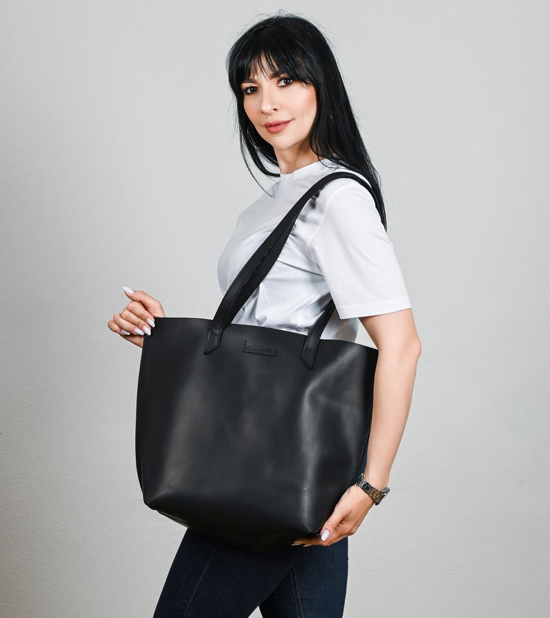 May include: A black leather tote bag with two shoulder straps. The bag is rectangular with a flat bottom and a minimalist design. The bag is held by a person wearing a white t-shirt and blue jeans.