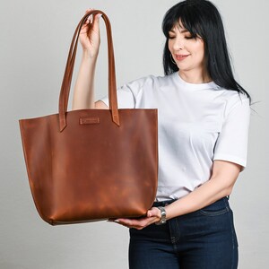 May include: A brown leather tote bag with long handles. The bag is held by a person. The bag has a rectangular shape with a wide opening and a small rectangular label near the top. The leather has a natural, textured appearance.
