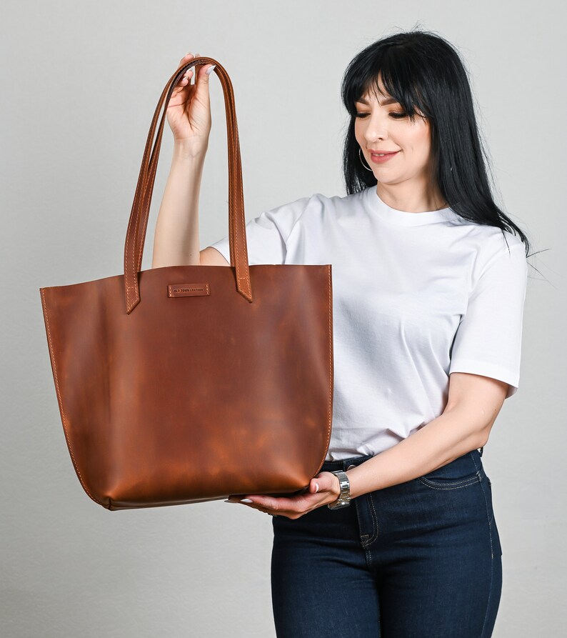 May include: A brown leather tote bag with long handles. The bag is held by a person. The bag has a rectangular shape with a wide opening and a small rectangular label near the top. The leather has a natural, textured appearance.