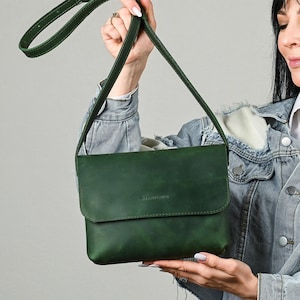 May include: A forest green crossbody bag with a rectangular flap closure and a long adjustable shoulder strap. The front of the bag features a small embossed logo that reads "BOLD TONE LEATHER" centered on the flap. The bag has a smooth finish.