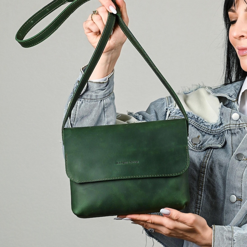 May include: A forest green crossbody bag with a rectangular flap closure and a long adjustable shoulder strap. The front of the bag features a small embossed logo that reads "BOLD TONE LEATHER" centered on the flap. The bag has a smooth finish.
