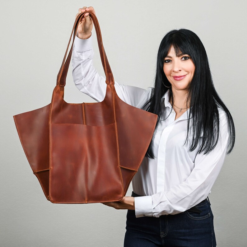 Pode incluir: Uma mulher segura uma grande bolsa tote de couro marrom com uma parte superior larga e aberta e um &uacute;nico bolso na frente. A bolsa tem uma forma relaxada e ligeiramente ca&iacute;da.