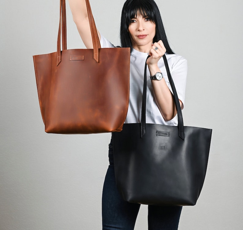 May include: Two leather tote bags, one in a rich brown and the other in black, are displayed. Both bags have long handles and a rectangular shape. The brown bag has a natural, textured appearance, while the black bag has a smooth finish. Both bags have a small rectangular tag.