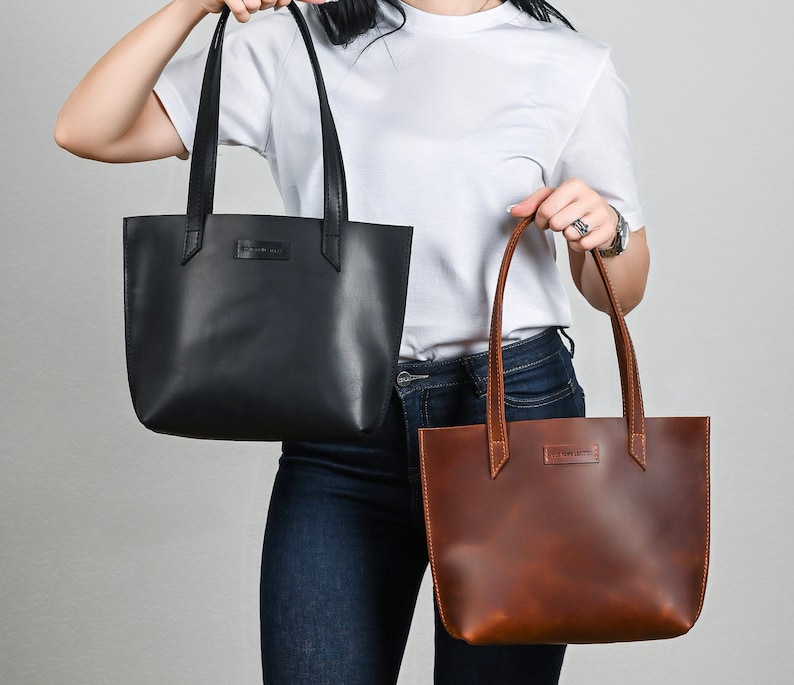 May include: Two leather tote bags, one black and one brown, are being held by a person wearing a white t-shirt and blue jeans.