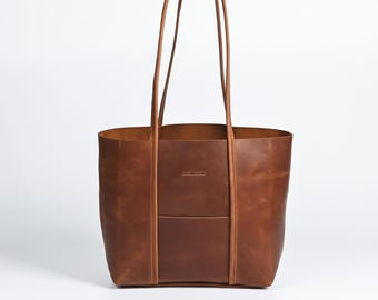 May include: A brown leather tote bag with long handles. The bag has a rectangular shape with a front pocket and a small logo. The leather has a natural, textured appearance.