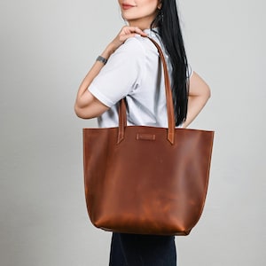 May include: A large, brown leather tote bag with long handles. The bag has a rectangular shape with a slightly rounded bottom. The leather appears to have a natural grain and texture. The handles are attached to the bag with reinforced stitching.