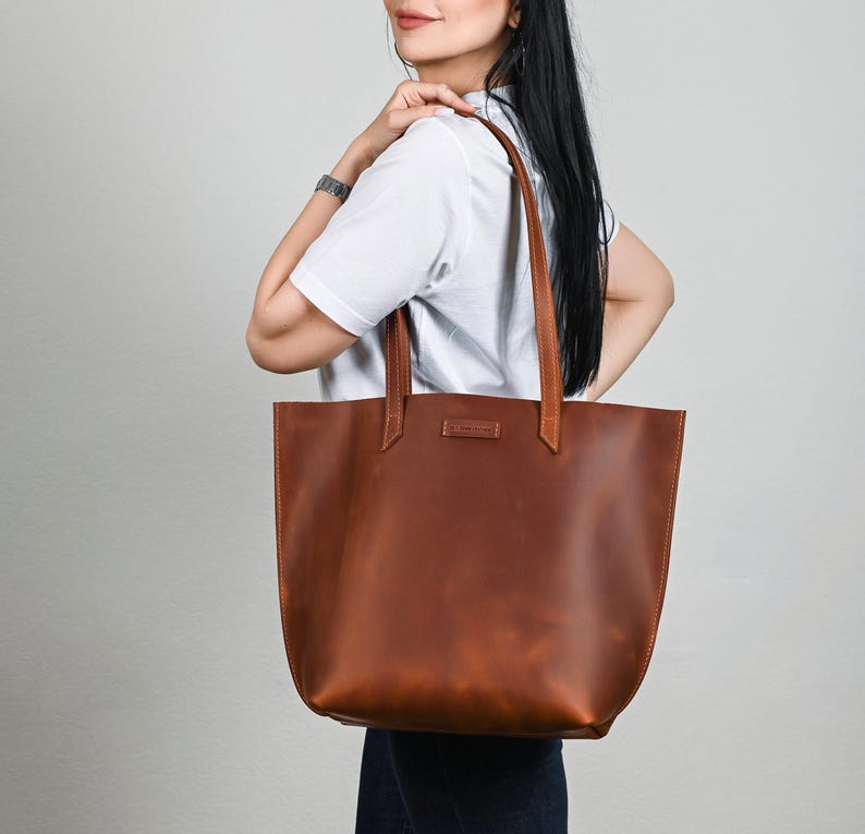 May include: A large, brown leather tote bag with long handles. The bag has a rectangular shape with a slightly rounded bottom. The leather appears to have a natural grain and texture. The handles are attached to the bag with reinforced stitching.