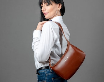 May include: A woman wearing a white button-down shirt and blue jeans is carrying a brown leather shoulder bag. The bag has a silver buckle and a silver logo that reads "A.P.C. Rue Madame".