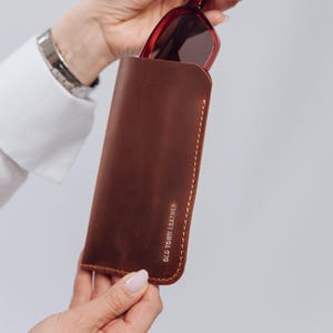 Może przedstawiać: Brązowe skórzane etui na okulary przeciwsłoneczne z wytłoczonym tekstem "OLD TOWN LEATHER" na boku. Etui jest zamknięte, a para czerwonych okularów przeciwsłonecznych wystaje częściowo z górnej części.