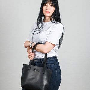 May include: A woman wearing a white t-shirt and blue jeans is holding a black leather tote bag.