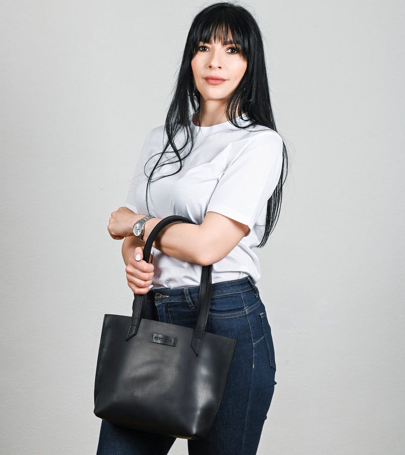 May include: A woman wearing a white t-shirt and blue jeans is holding a black leather tote bag.