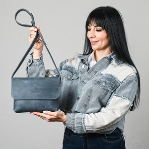 May include: A slate blue rectangular crossbody bag with a fold-over flap closure and a long adjustable shoulder strap. The bag features a smooth matte finish and a small embossed logo on the front flap, providing a clean and functional design.