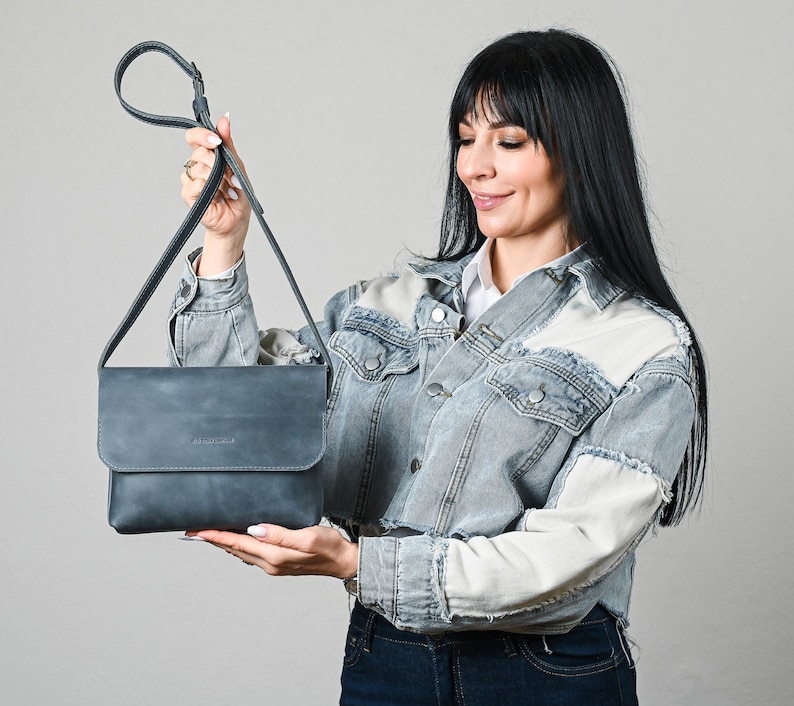 May include: A slate blue rectangular crossbody bag with a fold-over flap closure and a long adjustable shoulder strap. The bag features a smooth matte finish and a small embossed logo on the front flap, providing a clean and functional design.