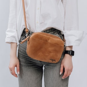 May include: A brown leather crossbody bag with a zipper closure and a strap. The bag has the text "Crafted for the modern nomad" printed on it.