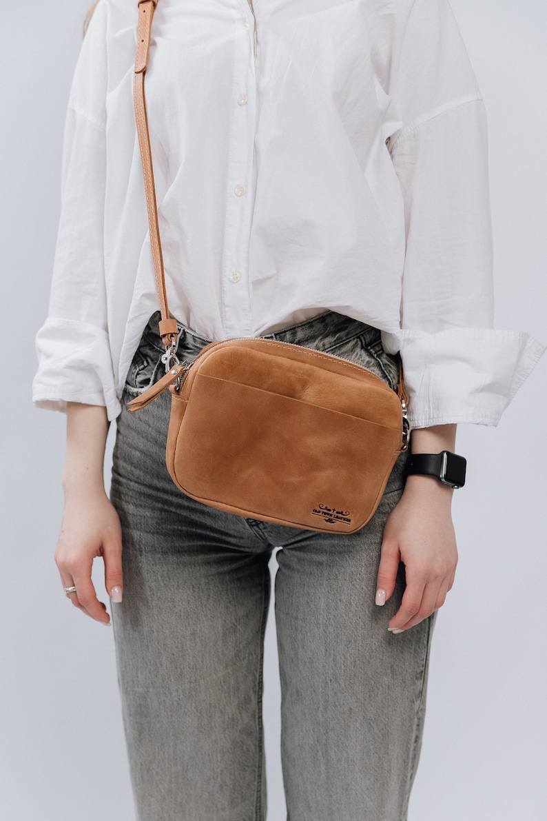 May include: A brown leather crossbody bag with a zipper closure and a strap. The bag has the text "Crafted for the modern nomad" printed on it.