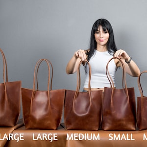 May include: Five brown leather tote bags of different sizes lined up on a table. The bags are labeled "Extra Large", "Large", "Medium", "Small", and "Mini".