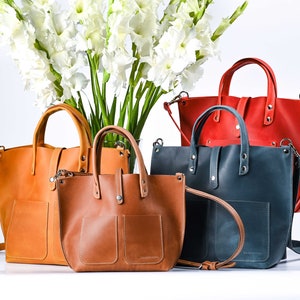 May include: Four leather tote bags in different colors and sizes. The bags have a top handle and a shoulder strap. The bags are arranged in a row on a white surface.