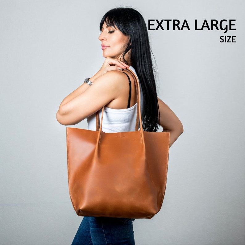 May include: A woman is holding a large brown leather tote bag. The bag has a simple design and is made of high-quality leather. The bag is labeled "EXTRA LARGE SIZE".