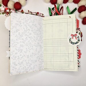 May include: Open journal with lined pages and a floral patterned page. A decorative Christmas wreath tag is attached. Red and white felt ball garland and Christmas ribbon accents the journal.