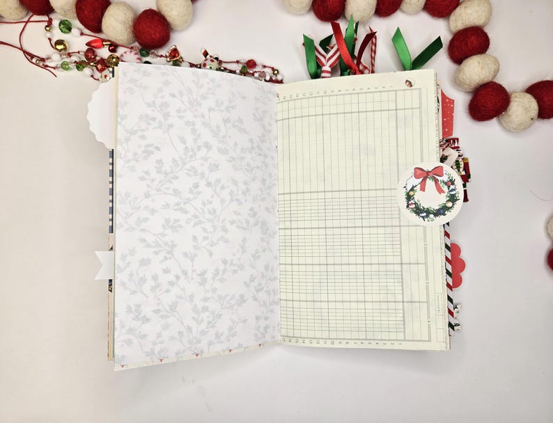May include: Open journal with lined pages and a floral patterned page. A decorative Christmas wreath tag is attached. Red and white felt ball garland and Christmas ribbon accents the journal.