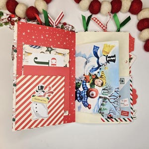 May include: Open Christmas-themed journal with red and white striped and star-patterned pages. Features a snowman illustration and a vintage winter scene. Includes pockets and decorative ribbons. A festive holiday craft.