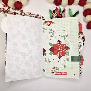 May include: Open journal with Christmas-themed pages. One page features a white floral pattern, while the other showcases a doily with a poinsettia design. The journal includes decorative ribbons and a "Comfort & Joy" label.