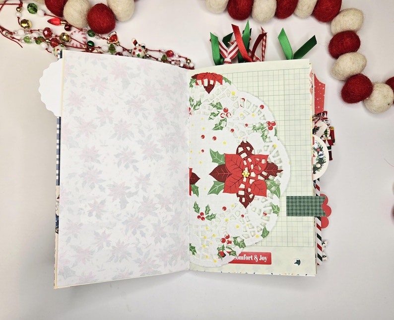 May include: Open journal with Christmas-themed pages. One page features a white floral pattern, while the other showcases a doily with a poinsettia design. The journal includes decorative ribbons and a "Comfort & Joy" label.