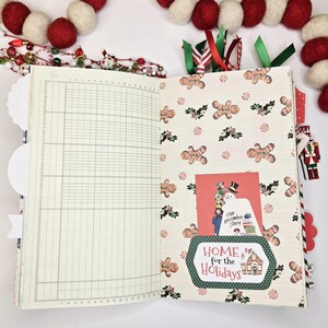May include: Open journal with lined pages and a Christmas-themed patterned page featuring gingerbread men and holly. A card with a snowman and the text "Home for the Holidays" is tucked inside. Red and white felt ball garland in the background.