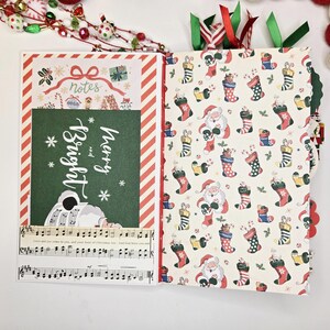 May include: Open Christmas-themed journal with festive patterned pages. One side features a green panel with "Merry and Bright" text and musical notes. The other side has a pattern of Santa Claus and stockings. Red and green ribbons mark the pages.