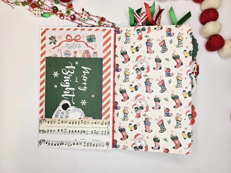 May include: Open Christmas-themed journal with festive patterned pages. One side features a green panel with "Merry and Bright" text and musical notes. The other side has a pattern of Santa Claus and stockings. Red and green ribbons mark the pages.