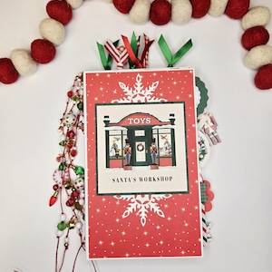 May include: A festive Christmas-themed scrapbook with a red cover featuring a snowflake design and the words "TOYS" and "SANTA'S WORKSHOP." The scrapbook is adorned with green, red, and white ribbons, and a garland of red and white felt balls.
