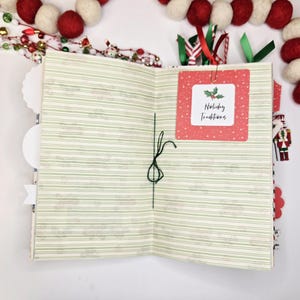 May include: Open journal with green striped pages and a red square label that reads "Holiday Traditions." The journal is decorated with red and white felt ball garland and festive ribbons. A small nutcracker figure is visible.