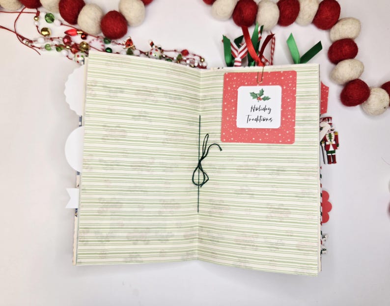 May include: Open journal with green striped pages and a red square label that reads "Holiday Traditions." The journal is decorated with red and white felt ball garland and festive ribbons. A small nutcracker figure is visible.