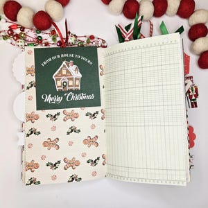 May include: Open journal with Christmas-themed pages. One page features a green card with a house illustration and the text "From Our House to Yours" and "Merry Christmas." The other page is lined. Festive decorations surround the journal.
