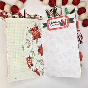 May include: Open journal with Christmas-themed pages. One page has graph paper, the other has a white floral pattern. A decorative doily with poinsettias and holly is on the left. The right page has a label that says "Christmas Cookie Crew". Ribbons and a garland are in the background.