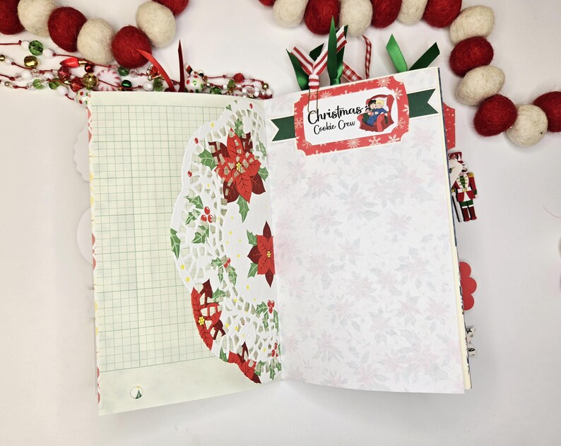May include: Open journal with Christmas-themed pages. One page has graph paper, the other has a white floral pattern. A decorative doily with poinsettias and holly is on the left. The right page has a label that says "Christmas Cookie Crew". Ribbons and a garland are in the background.