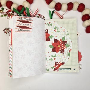 May include: Open journal with decorative pages. One page features a poinsettia design on a doily. The other page has text that reads "Let's Make It December To Remember". Red, green, and white ribbon bookmarks are visible.