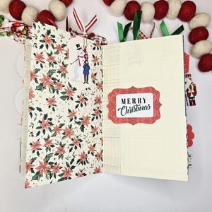 May include: Open journal with pages featuring poinsettia floral patterns and lined paper. A sticker with a snowman and child is on the left page. The right page has a "Merry Christmas" sticker. Red and white felt ball garland decorates the background.